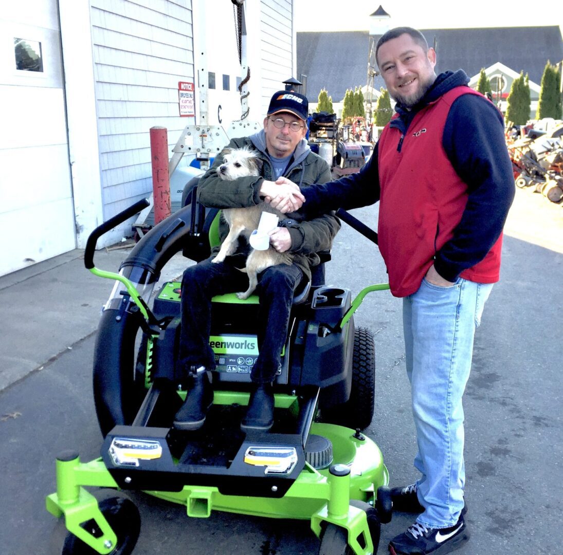 A man holding a teddy bear next to a person in a green lawn mower.